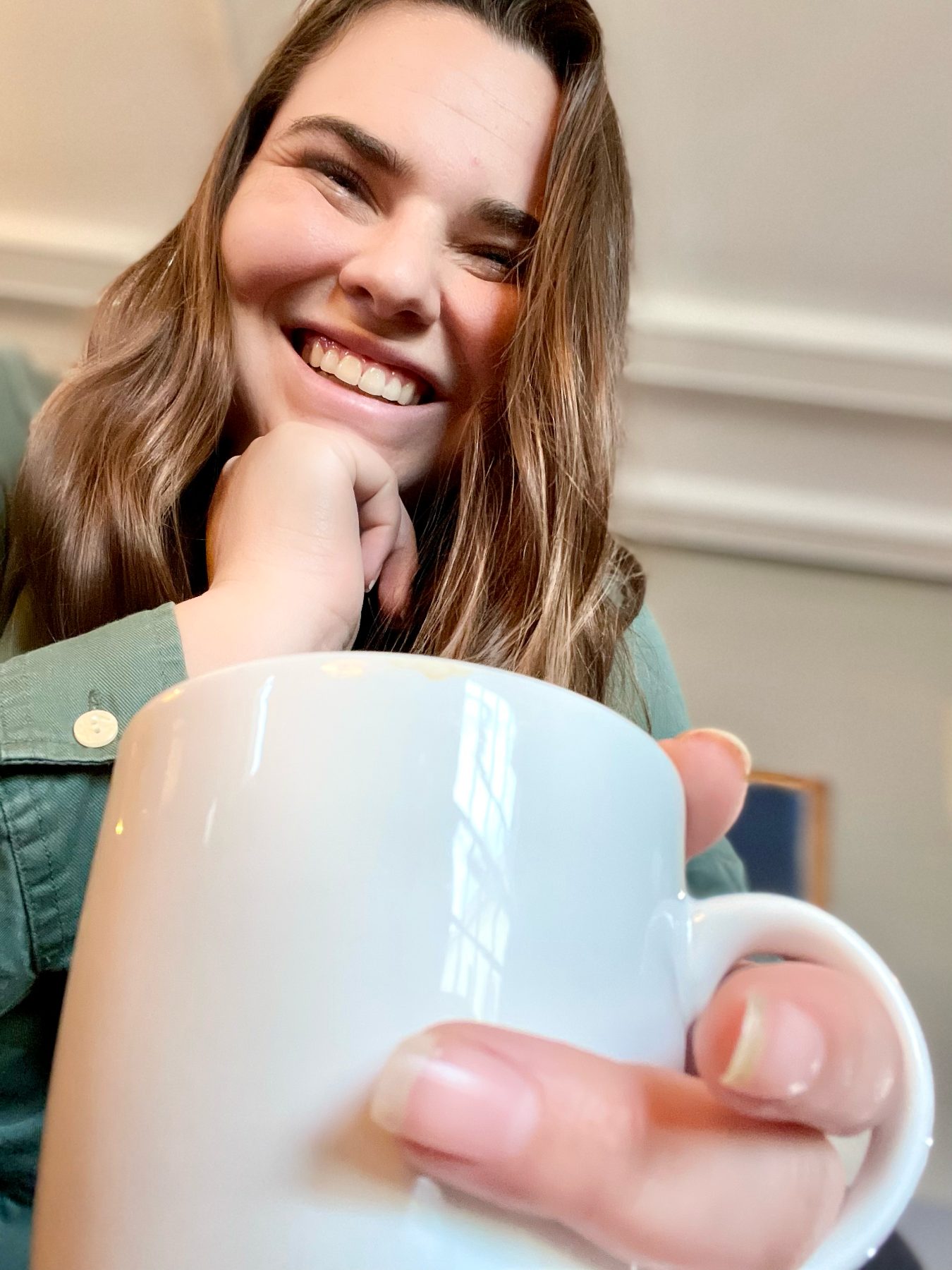 Andrea smiling with her chin on her hand, holding a white coffee mug toward the camera